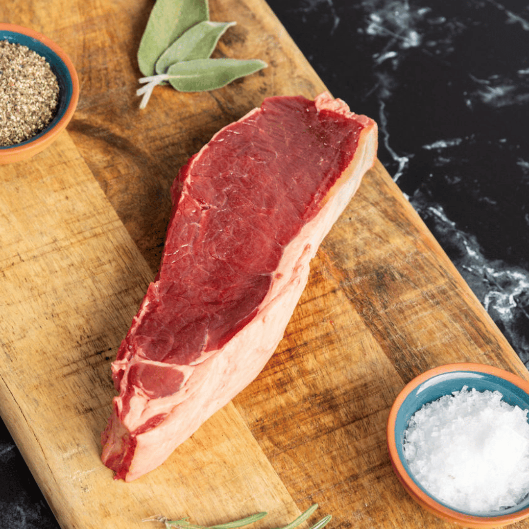 Bison New York strip steak uncooked on a wooden cutting board being prepared with salt, pepper, garlic and sage.