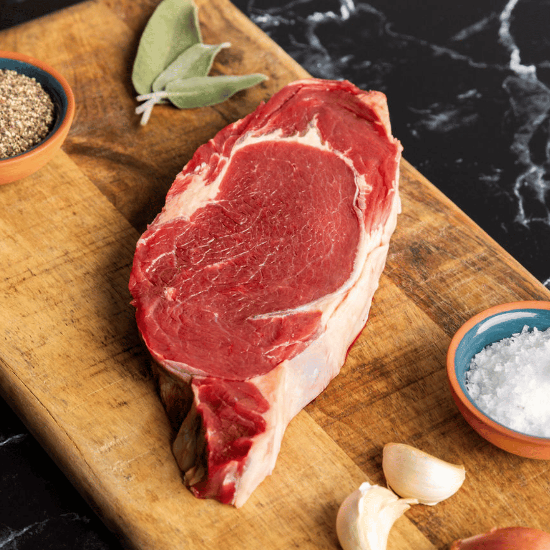Bison ribeye steak on wooden cutting board being prepared with salt, pepper, garlic and sage.