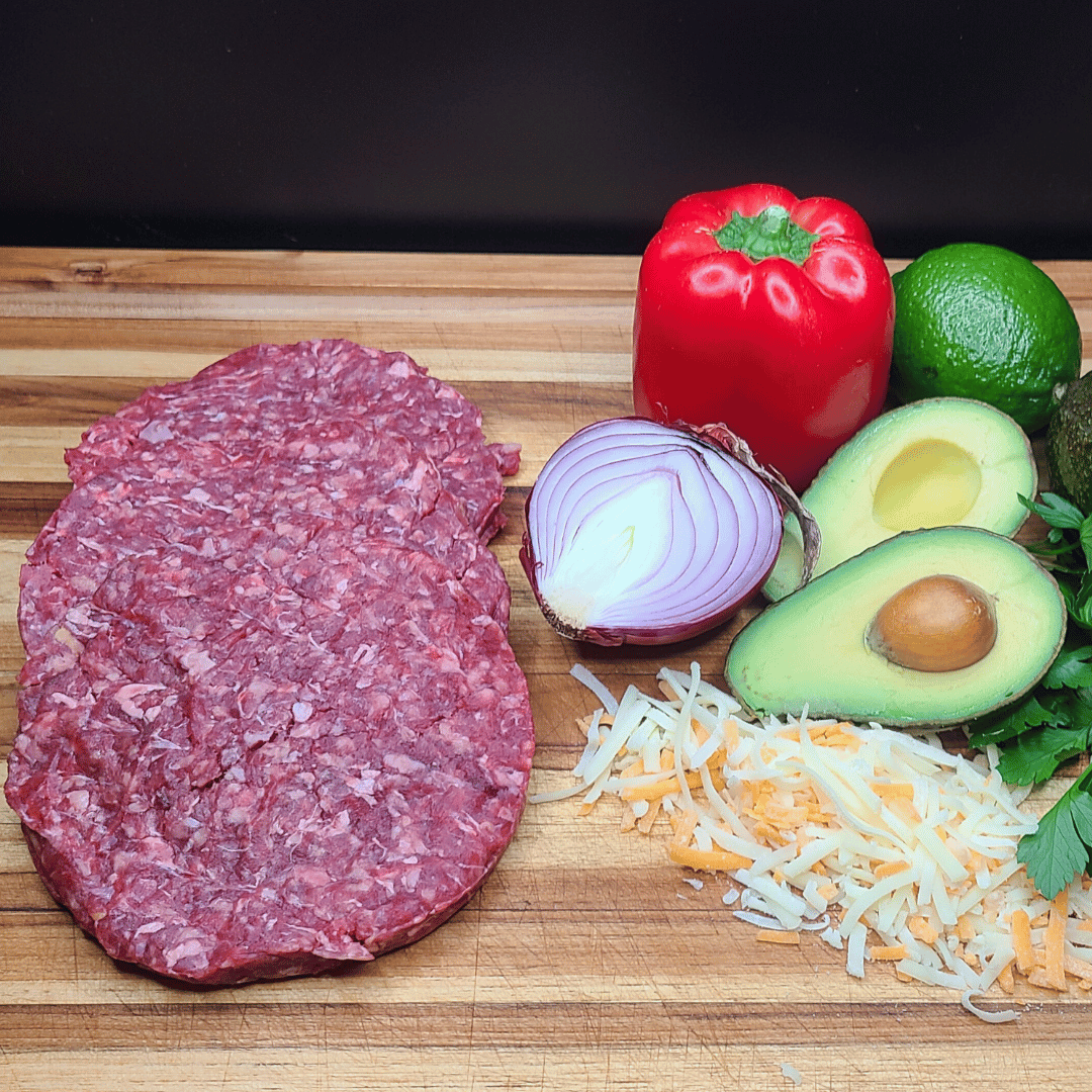 Ground bison burger on a wooden cutting board being prepared with fresh veggies and shredded cheese.