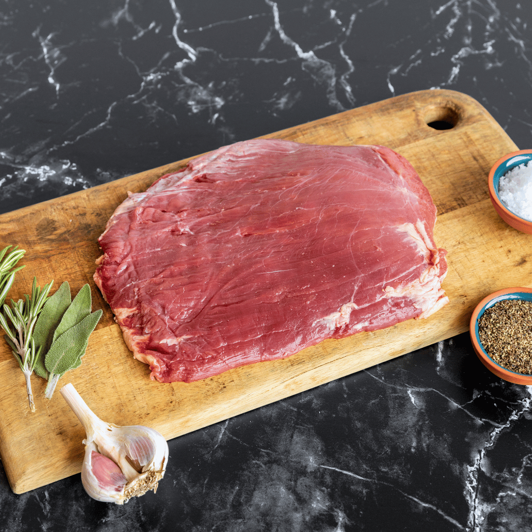 Bison flank steak on a wooden cutting board being prepared with salt, pepper, garlic, rosemary and sage.