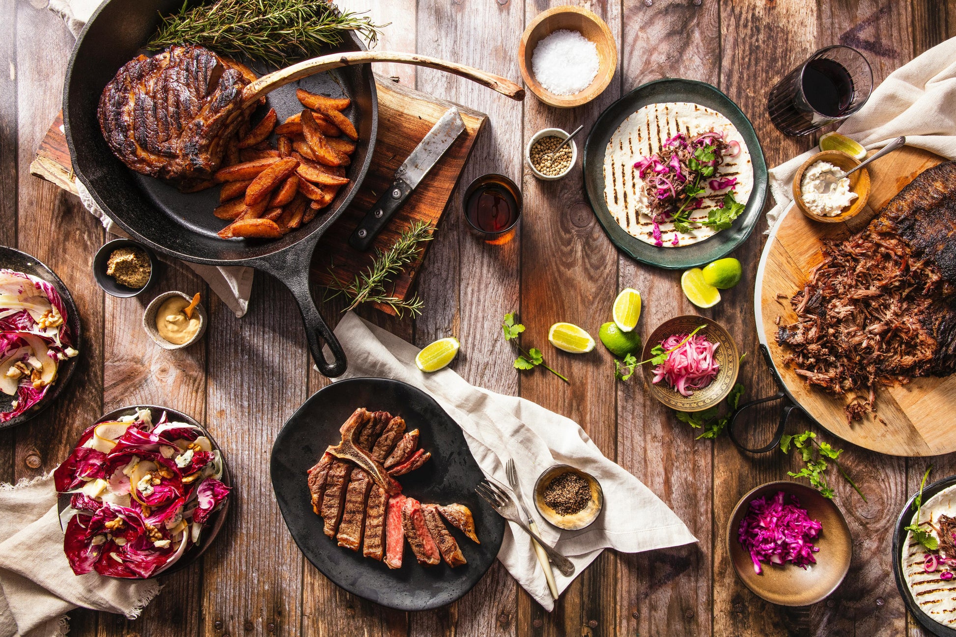 Wood table spread with variety of prepared grilled buffalo, veggies, tortillas, limes, sauces and iced tea.